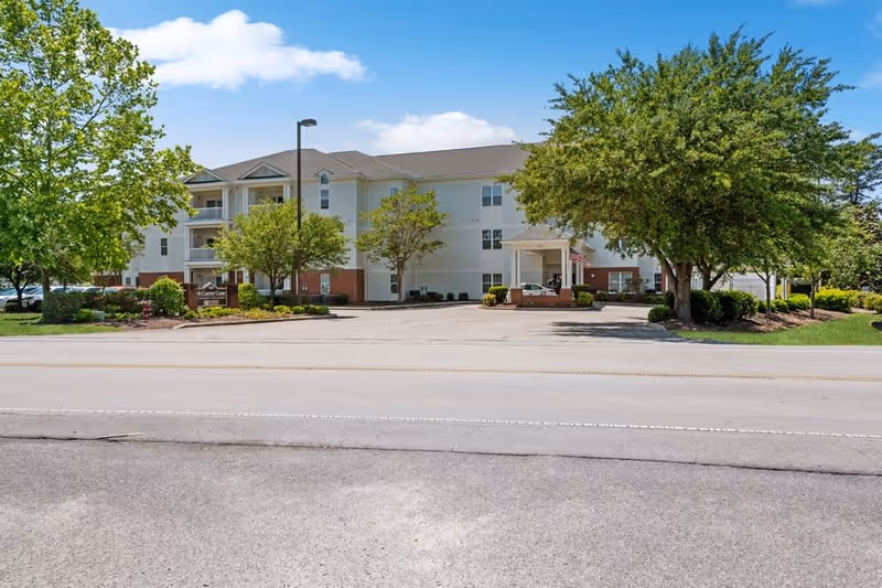 Exterior view of McCarthy Court, a three-story senior living facility with white and brick facade, surrounded by trees and landscaping under a blue sky with some clouds. There is a driveway and parking area in front of the building.