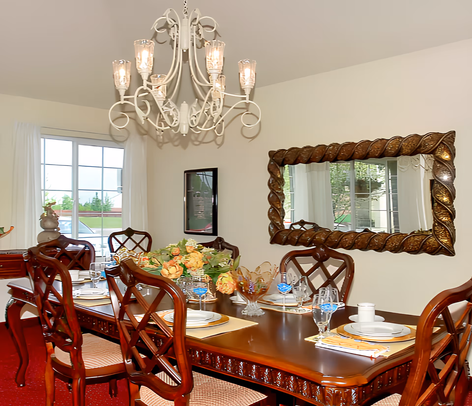 A formal dining room with a polished wooden table set for six people. The table is adorned with plates, glasses, napkins, and a floral centerpiece. There is a large decorative mirror on the wall and a white chandelier hanging from the ceiling. A window with white curtains allows natural light into the room.