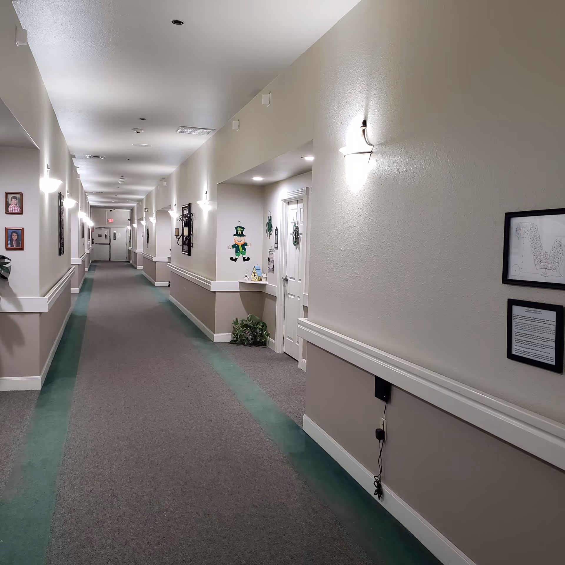 A long, well-lit hallway in a senior living facility with beige walls and green and gray carpet. The hallway has wall-mounted lights, framed pictures, and decorations including a leprechaun figure near a door. There are handrails along the walls and some plants placed near the doors.