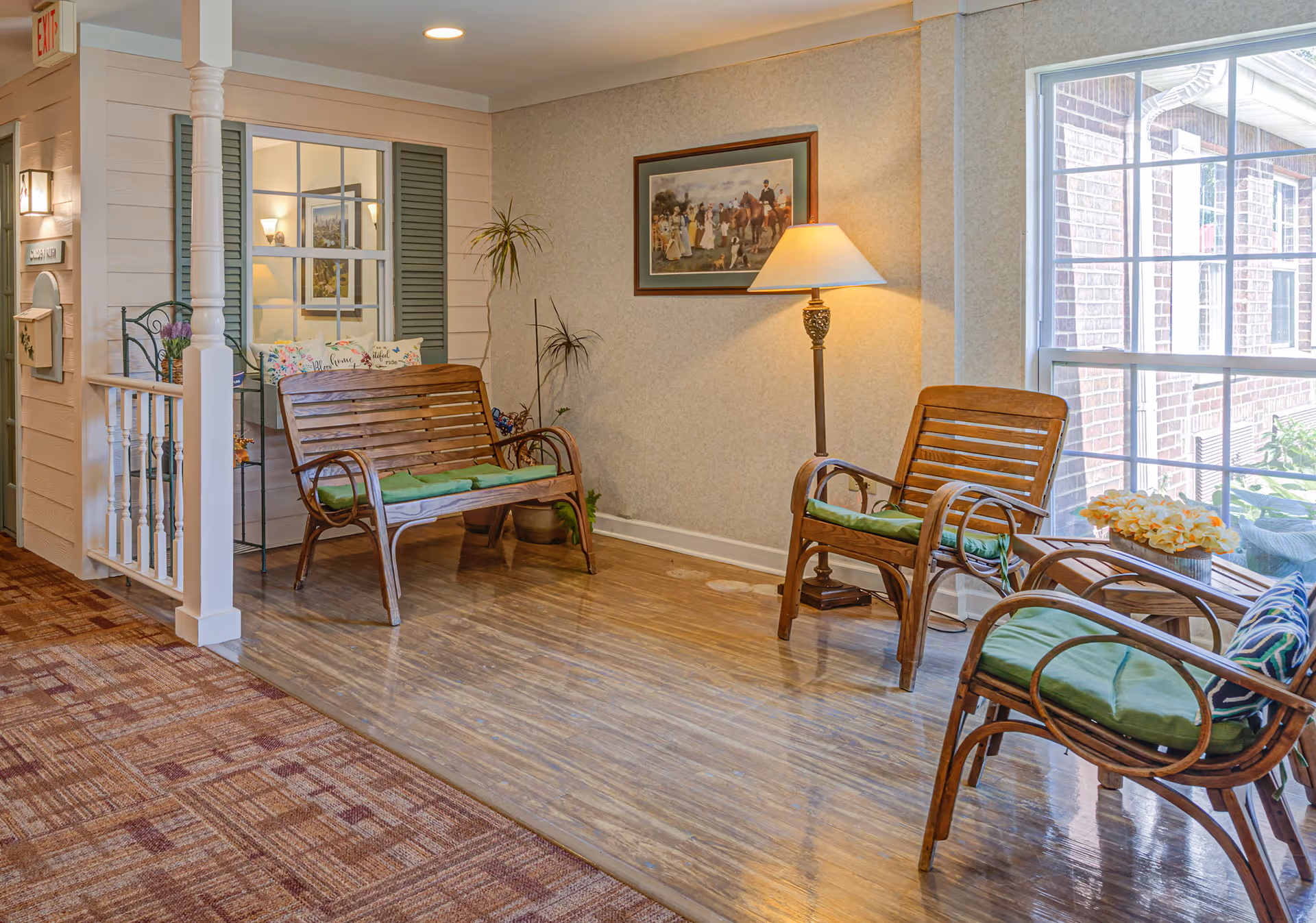A cozy seating area in a senior living facility with wooden chairs and a bench featuring green cushions. There is a floor lamp beside the bench, a framed painting on the wall, a large window letting in natural light, and some decorative plants and flowers. The floor transitions from carpet to wood, and there is a small railing with white spindles near the entrance.