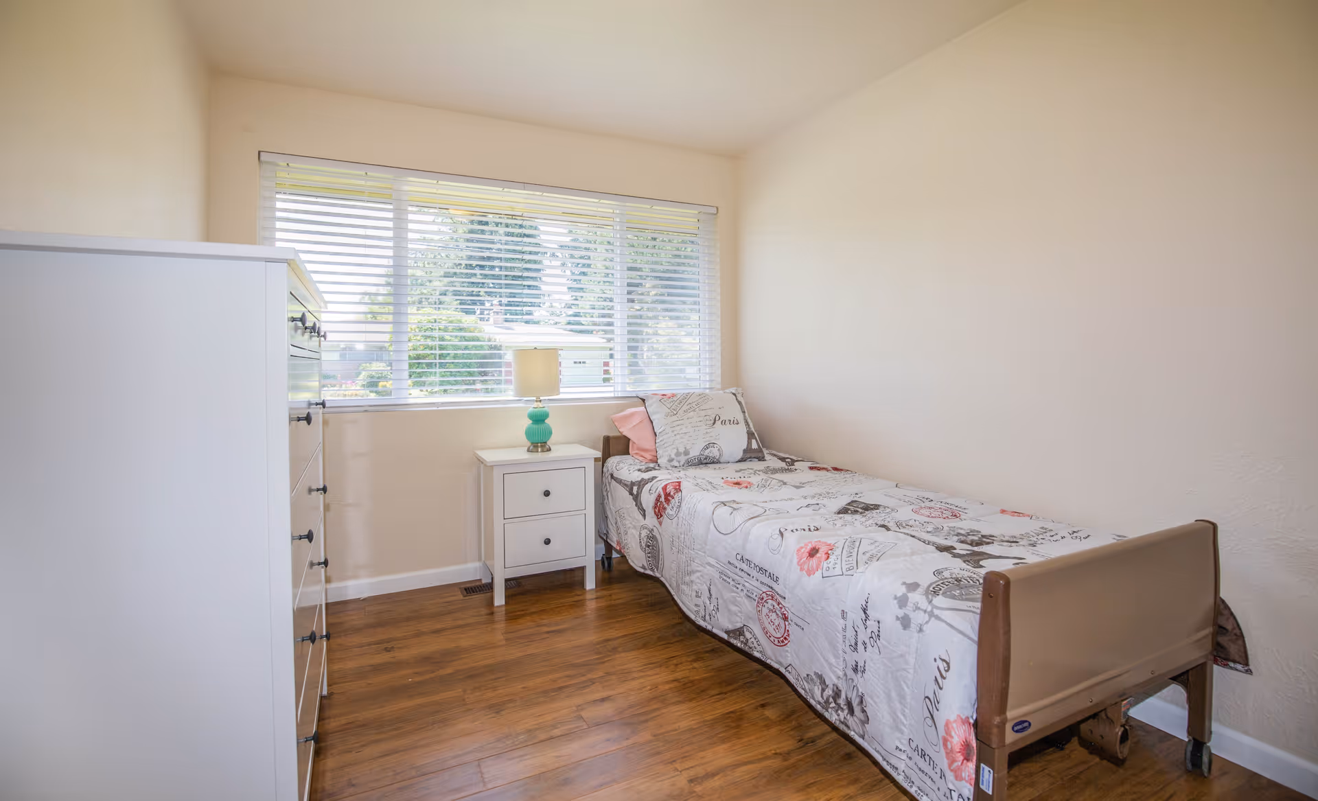 A small bedroom with a single bed covered in a Paris-themed quilt, a white nightstand with a turquoise lamp, a white dresser, and a large window with horizontal blinds letting in natural light.