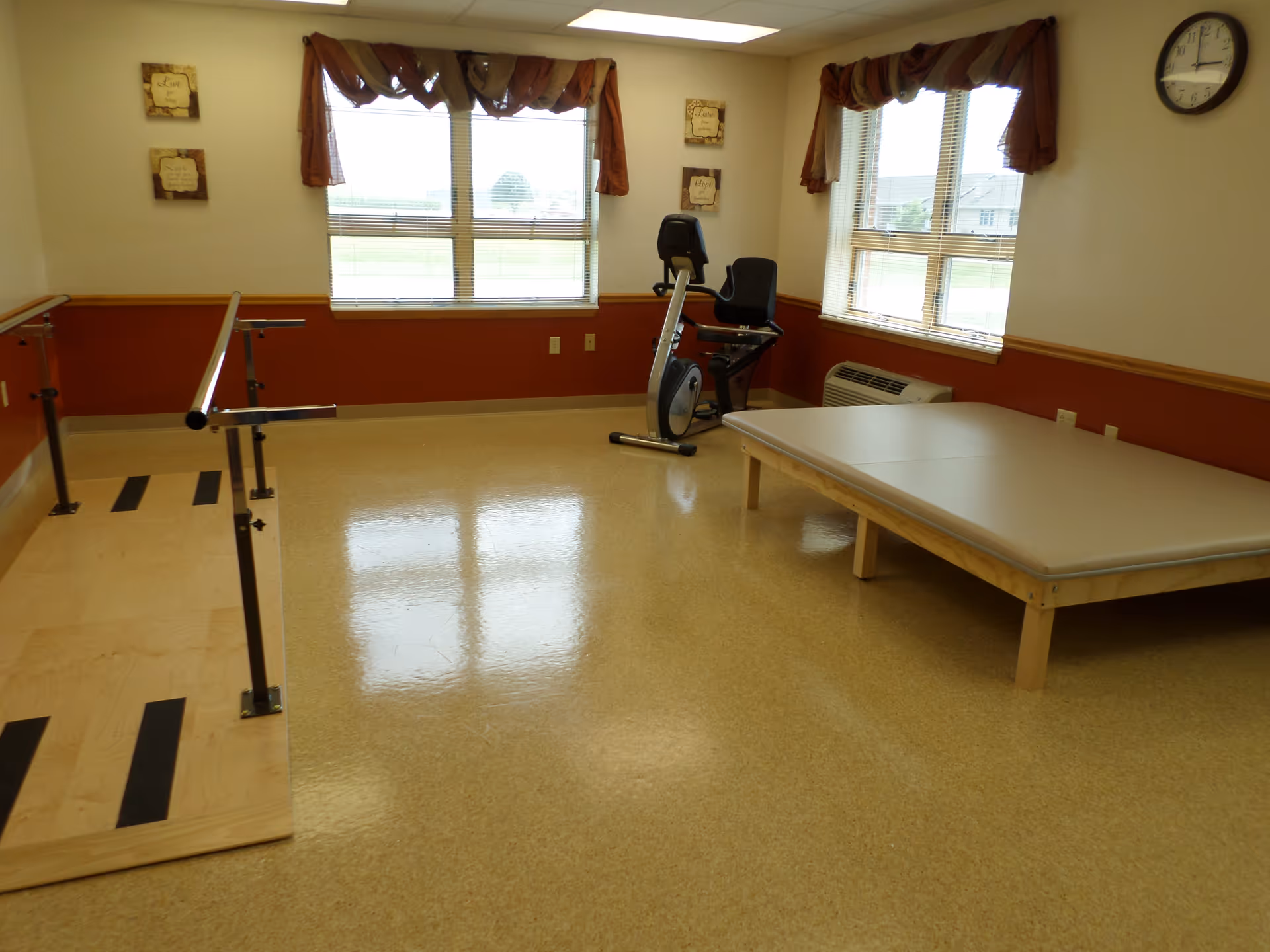 A rehabilitation room with a wooden parallel bar setup for walking exercises, a padded therapy table, and a stationary exercise bike. The room has two windows with brown valances, beige walls with a brown lower half, and a wall clock.
