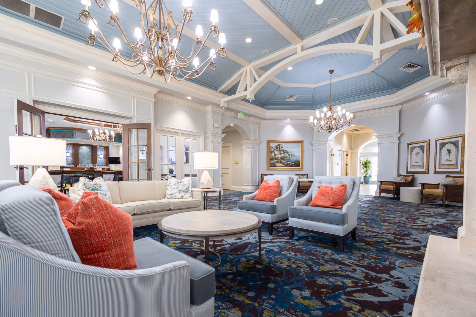 Bright, elegant senior living lobby with upholstered chairs and sofas around a round coffee table, ornate chandeliers, and a high blue-paneled ceiling.