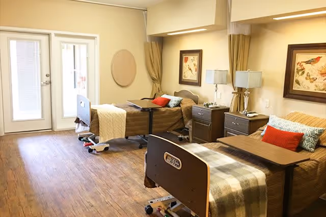 Shared senior living bedroom with two adjustable beds, nightstands, lamps, and patio doors.