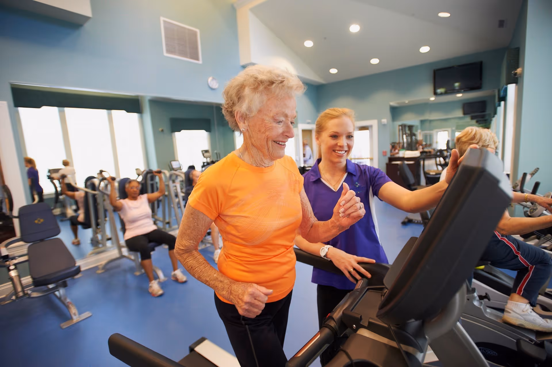 An elderly woman in an orange shirt is walking on a treadmill while a younger woman in a purple shirt assists her in a fitness room. Other elderly individuals are exercising on various gym equipment in the background. The room has blue floors, light blue walls, and large windows letting in natural light.