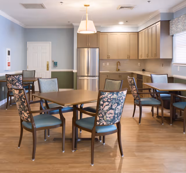 A bright and clean dining area with several wooden tables and chairs featuring floral upholstery. The room includes a modern kitchen in the background with beige cabinets, a stainless steel refrigerator, a gold faucet, and a window with a floral valance. The walls are painted light blue with a green wainscoting and white trim.