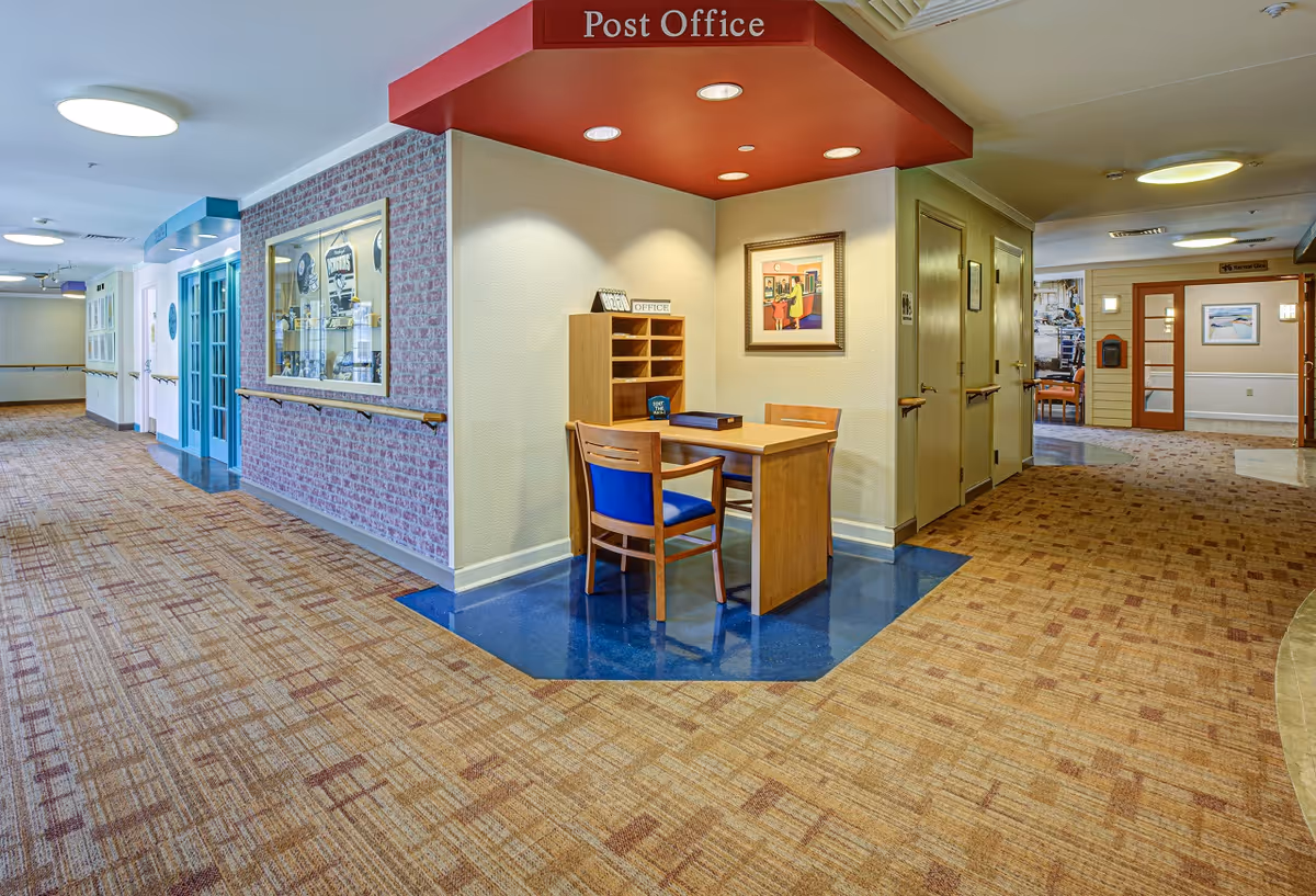A well-lit interior hallway of a memory care facility featuring a small 'Post Office' desk with chairs under a red canopy.