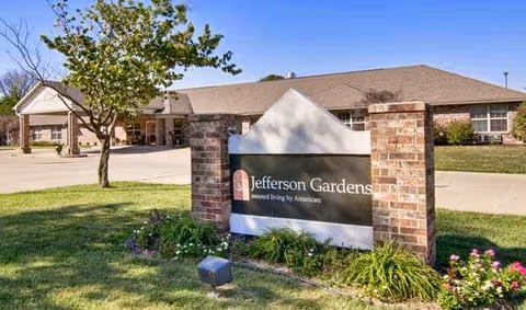 Exterior view of Jefferson Gardens Senior Living facility with a brick sign in the foreground displaying the facility's name. The building is single-story with a sloped roof, surrounded by a lawn, trees, and a driveway.