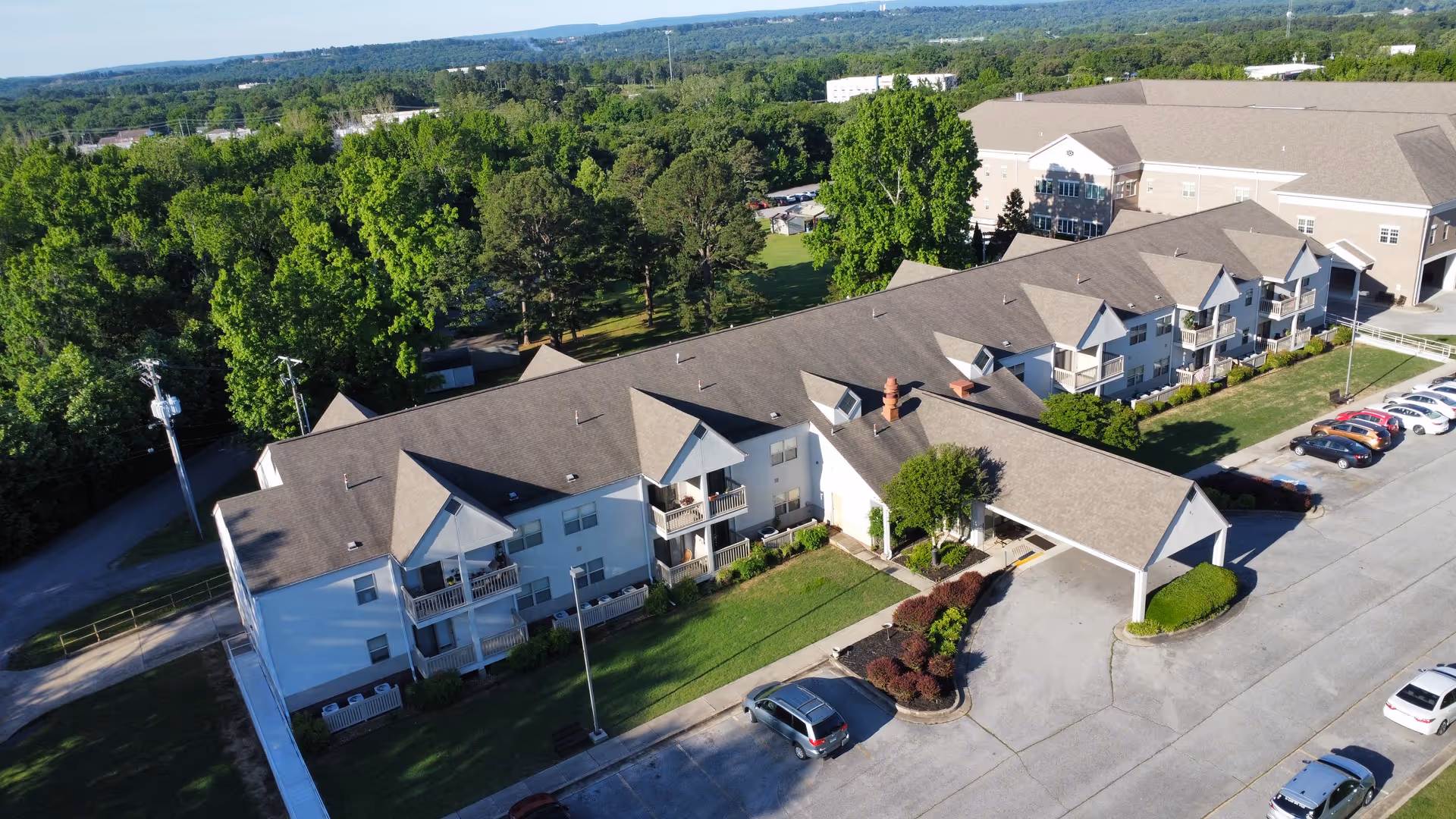 Aerial view of the Wildewood Independent Living complex showing connected apartment buildings with balconies, a covered drive-up entrance, parking lot, and surrounding trees.