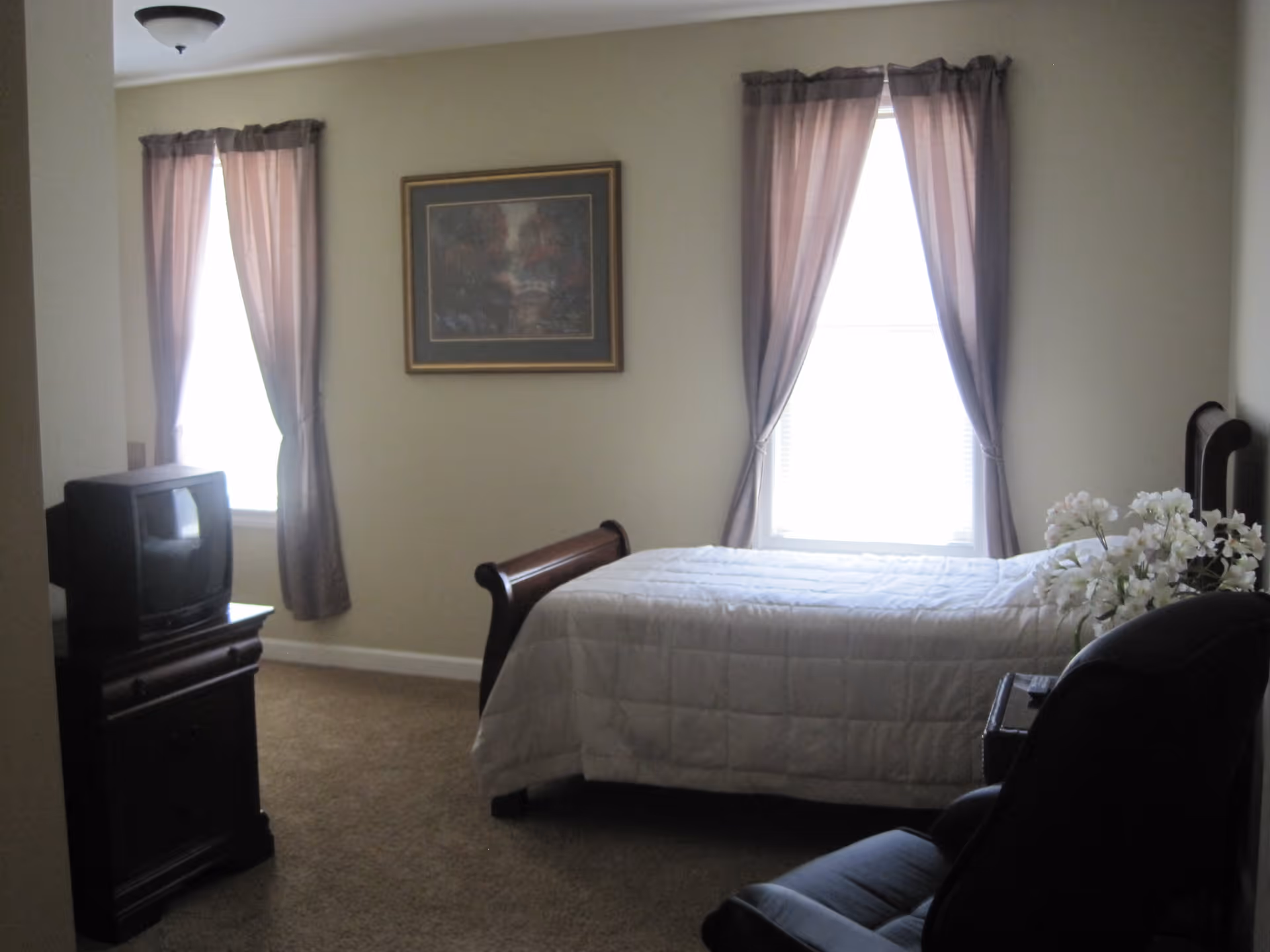 A bedroom with a single bed covered with a white quilt, two windows with sheer curtains, a wooden TV stand with an old-style CRT television, a framed painting on the wall, a dark armchair, and a small table with a vase of white flowers.