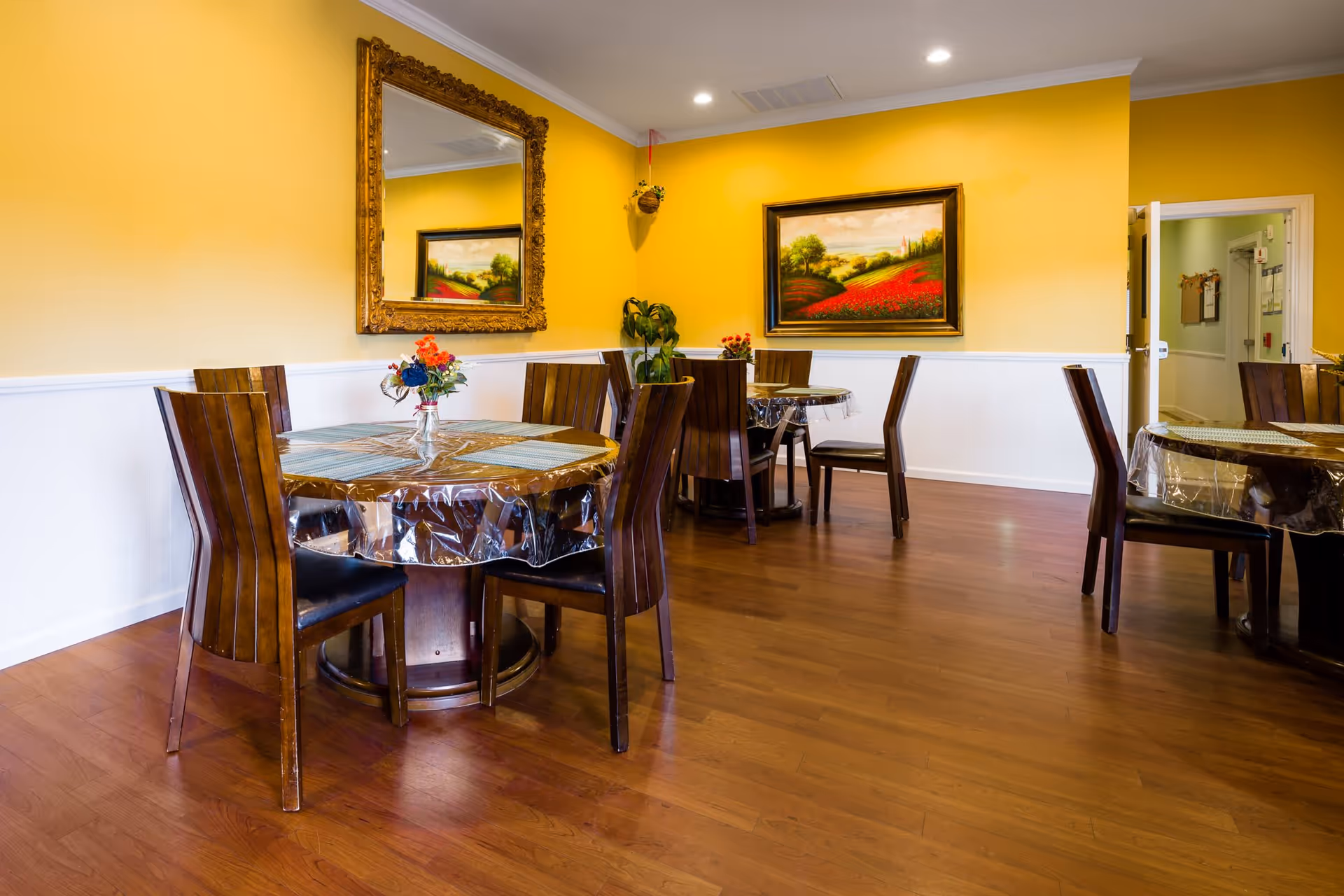 Dining room with round tables covered in plastic, wooden chairs, hardwood floors, and yellow walls adorned with framed paintings and a mirror.