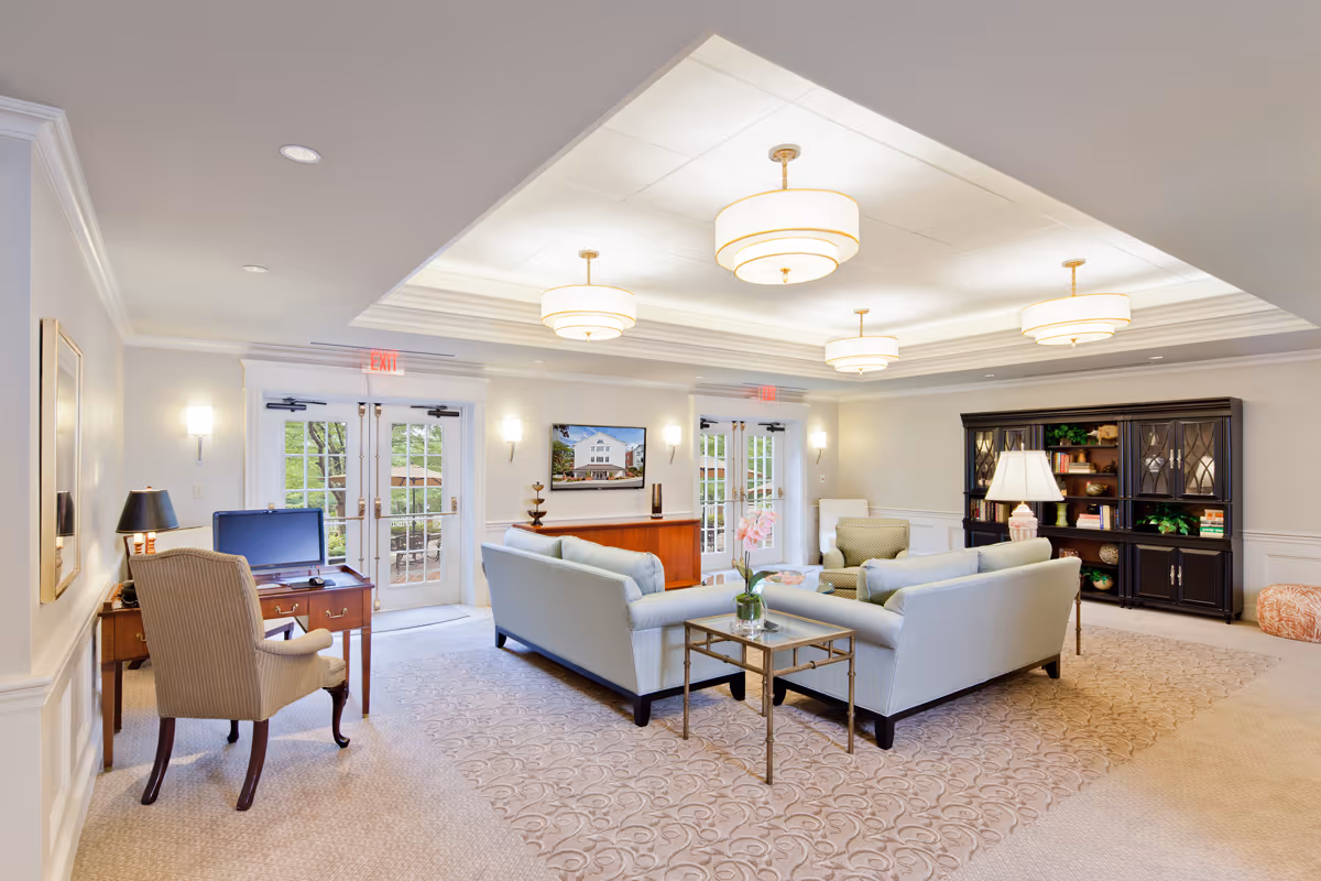 A bright and spacious living room area in Belmont Manor Nursing & Rehabilitation Center featuring light blue sofas arranged around a glass coffee table with a flower vase. The room has beige carpet with a patterned area, a dark wooden bookshelf with books and plants, a desk with a computer and chair, and multiple ceiling lights. French doors with glass panes lead outside, allowing natural light to fill the room.