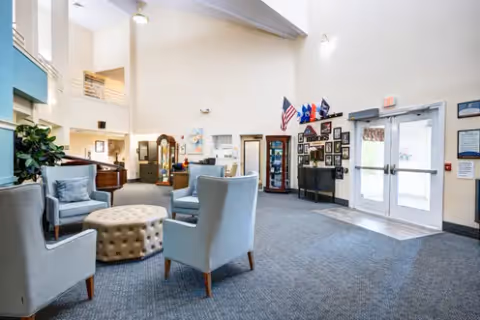 A spacious and well-lit living room area in a senior living facility with four light blue armchairs arranged around a round tufted ottoman. The room has high ceilings, a carpeted floor, and a glass door entrance. There are flags displayed on the wall, framed pictures, a grandfather clock, and a piano in the background.