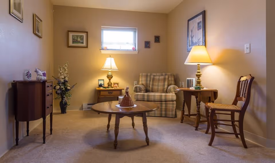 A cozy living room with beige walls and carpet, featuring a plaid armchair, a round wooden coffee table with a decorative item on it, two wooden side tables each with a lamp, a wooden chair with a woven seat, a small cabinet, and framed pictures on the walls. A small window lets in natural light.