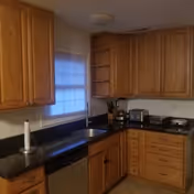 Interior view of a kitchen in an assisted living facility featuring wooden cabinets, a black countertop, a stainless steel dishwasher, a window with blinds, and various small kitchen appliances including a coffee maker and toaster.