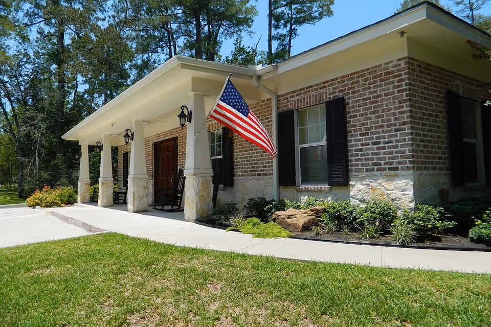 Exterior view of a single-story brick building with a covered porch supported by columns. The porch has two rocking chairs and an American flag mounted on one of the columns. There are windows with black shutters and a well-maintained lawn and garden beds surrounding the building.