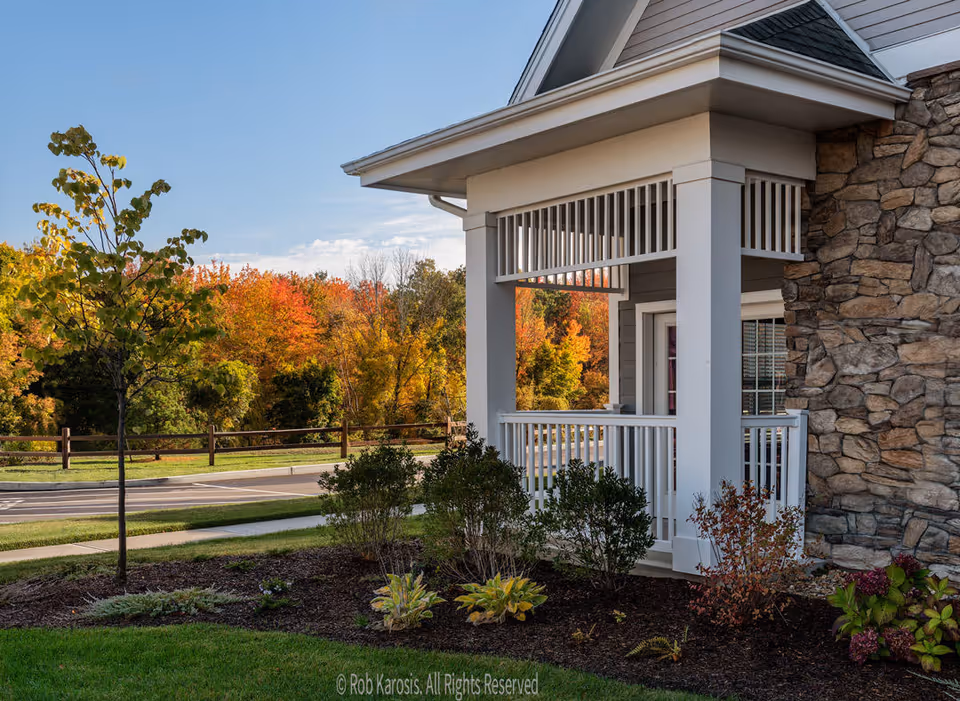 Exterior view of a retirement community building corner with stone and siding walls, a white porch railing, and landscaped bushes and plants in the foreground. Trees with autumn foliage and a clear blue sky are visible in the background.