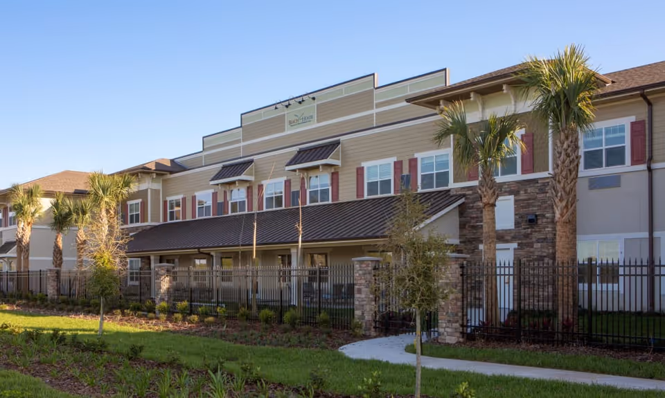Exterior view of a multi-story assisted living facility named Beach House Assisted Living & Memory Care, featuring beige and stone facade, multiple windows with red shutters, palm trees, a black metal fence, and a landscaped lawn under a clear blue sky.