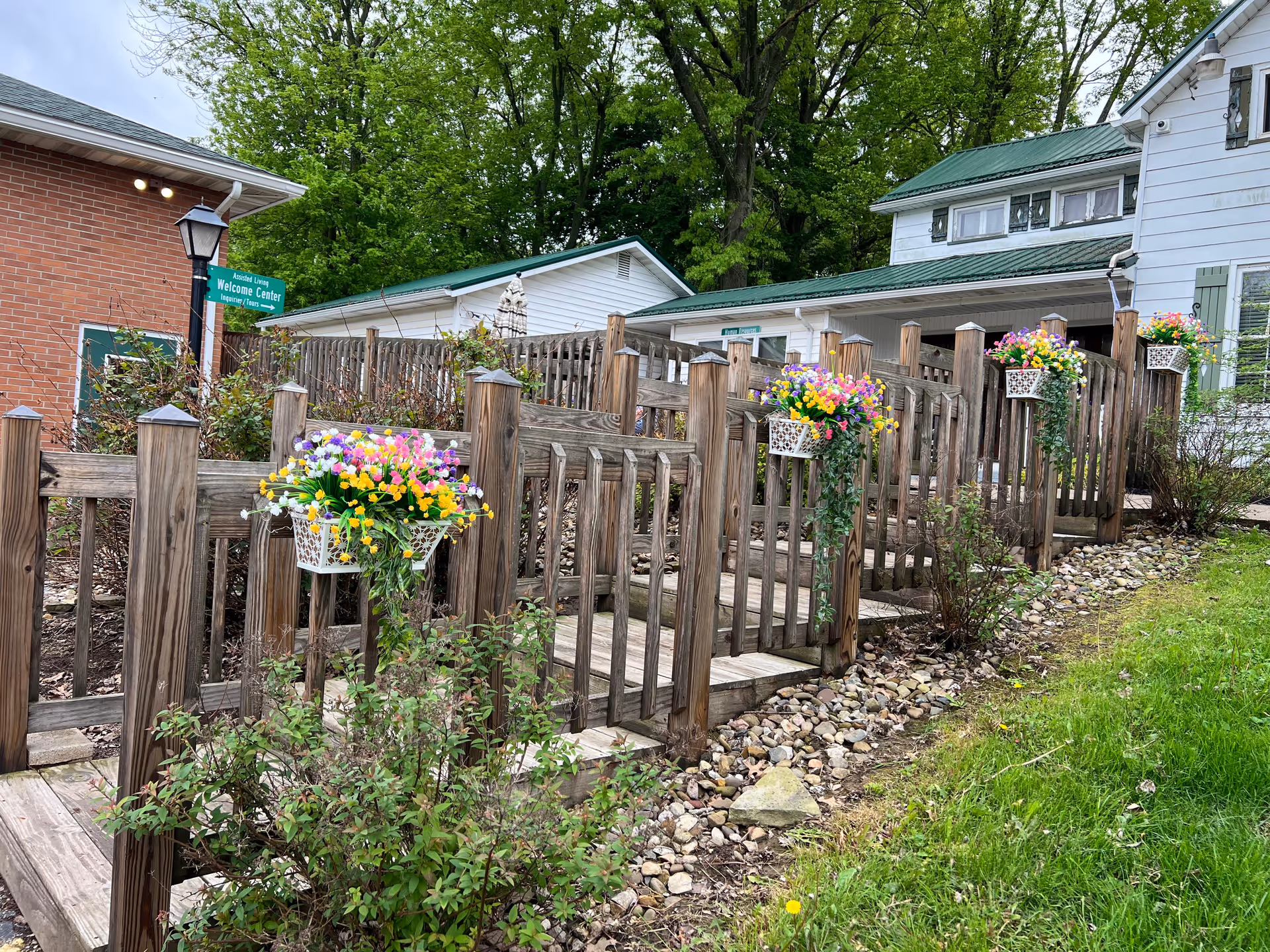Outdoor wooden ramp with handrails decorated with hanging baskets of colorful flowers leading to a building with white siding and green roof, surrounded by greenery and trees.