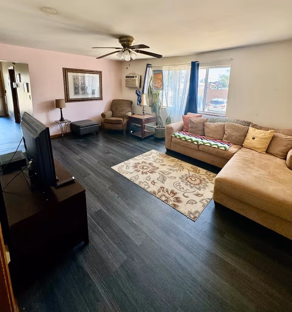 A cozy living room with a beige sectional sofa adorned with multiple cushions and a colorful throw blanket. There is a patterned area rug on the dark wood floor. A brown armchair and ottoman are placed near a window with sheer and dark blue curtains. A ceiling fan with lights hangs from the ceiling. A TV is positioned on a dark wooden stand facing the sofa. A framed picture hangs on the light pink wall, and a potted plant sits near the window.