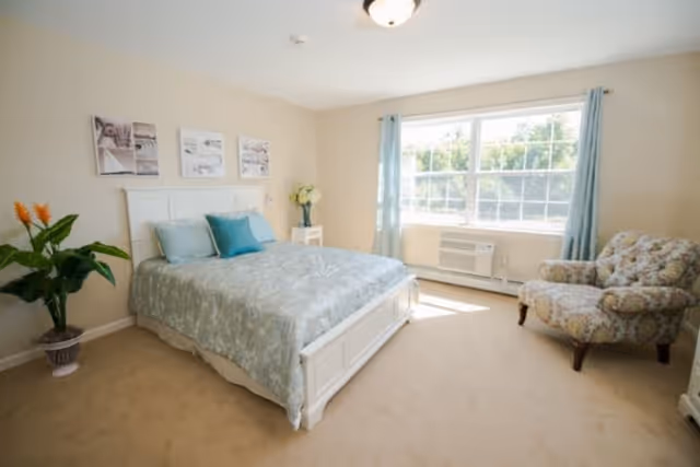 A bright bedroom with a large window letting in natural light. The room features a white bed frame with a light blue patterned bedspread and matching pillows. There is a floral armchair near the window, a small white nightstand with a vase of flowers, and a potted plant in the corner. The walls are light-colored, and three framed pictures hang above the bed.