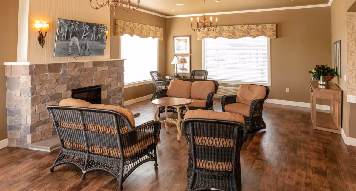 A cozy living room with wicker furniture including a loveseat, three armchairs, and a round wooden coffee table. The room features a stone fireplace with a flat-screen TV mounted above it displaying a black and white dance scene. Two large windows with floral valances allow natural light to fill the space. There are two chandeliers hanging from the ceiling and a wooden console table with a vase of flowers on the right side of the room.