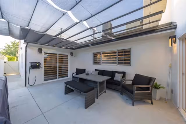Outdoor patio area with a shaded canopy overhead, featuring a black cushioned seating set including a sofa, armchair, bench, and a rectangular table. The patio is adjacent to a building with sliding glass doors and a window with horizontal bars.
