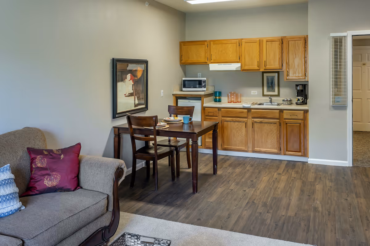 Interior view of a senior living facility unit at Lamar Court featuring a small kitchen area with wooden cabinets, a microwave, a coffee maker, and a sink. Adjacent to the kitchen is a wooden dining table set for two with plates and mugs. A beige sofa with decorative pillows is partially visible on the left side. The floor is a combination of wood and carpet, and there is a framed artwork on the wall above the dining table.