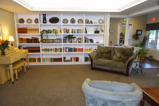 A cozy living room area with a beige piano on the left, a large white bookshelf filled with books and decorative items in the center, a green loveseat with patterned cushions to the right, and a light blue armchair in the foreground. There are plants and lamps adding warmth to the space.