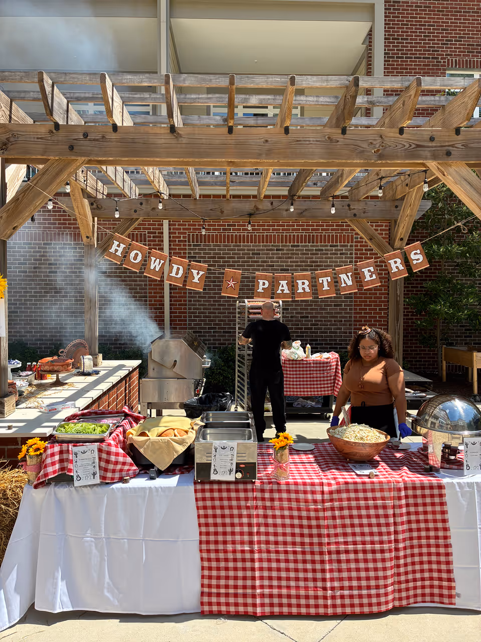 Outdoor barbecue buffet setup under a wooden pergola with a 'HOWDY PARTNERS' banner, red-checked tablecloths, serving dishes, and two staff members.