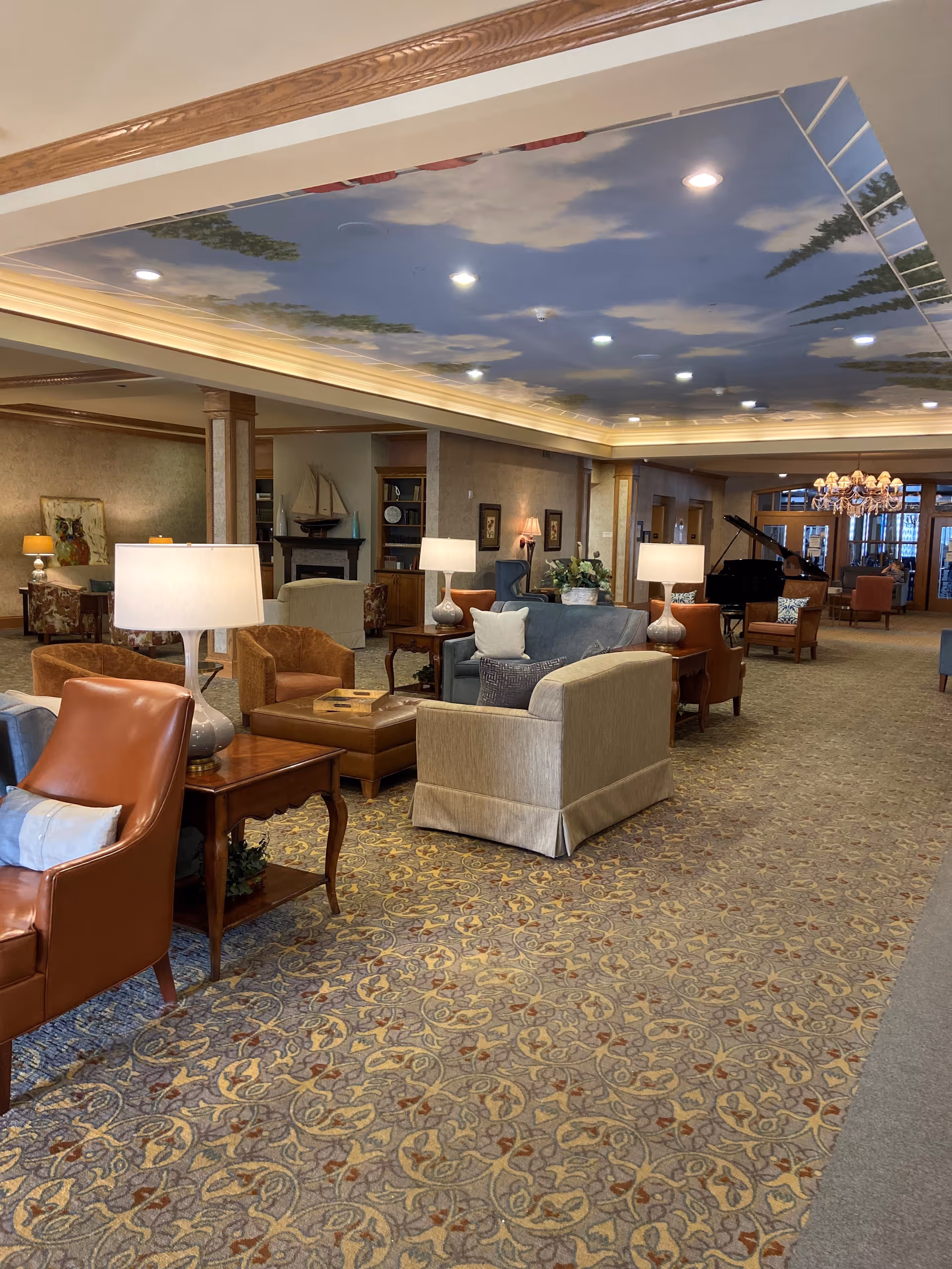 A spacious and elegantly furnished senior living facility common area with multiple seating arrangements including armchairs and sofas. The ceiling is painted with a sky mural featuring clouds and trees, and there are several lamps providing warm lighting. A grand piano and chandelier are visible in the background, along with decorative elements such as framed artwork and a model sailboat on a mantel.