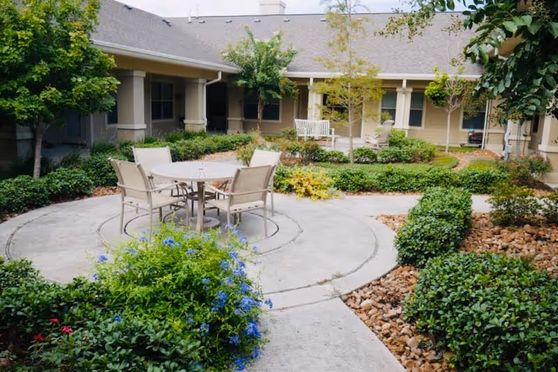 Outdoor courtyard area with a round table and four chairs on a concrete patio surrounded by green bushes, small trees, and flowering plants. The courtyard is enclosed by a beige building with windows and a covered walkway.