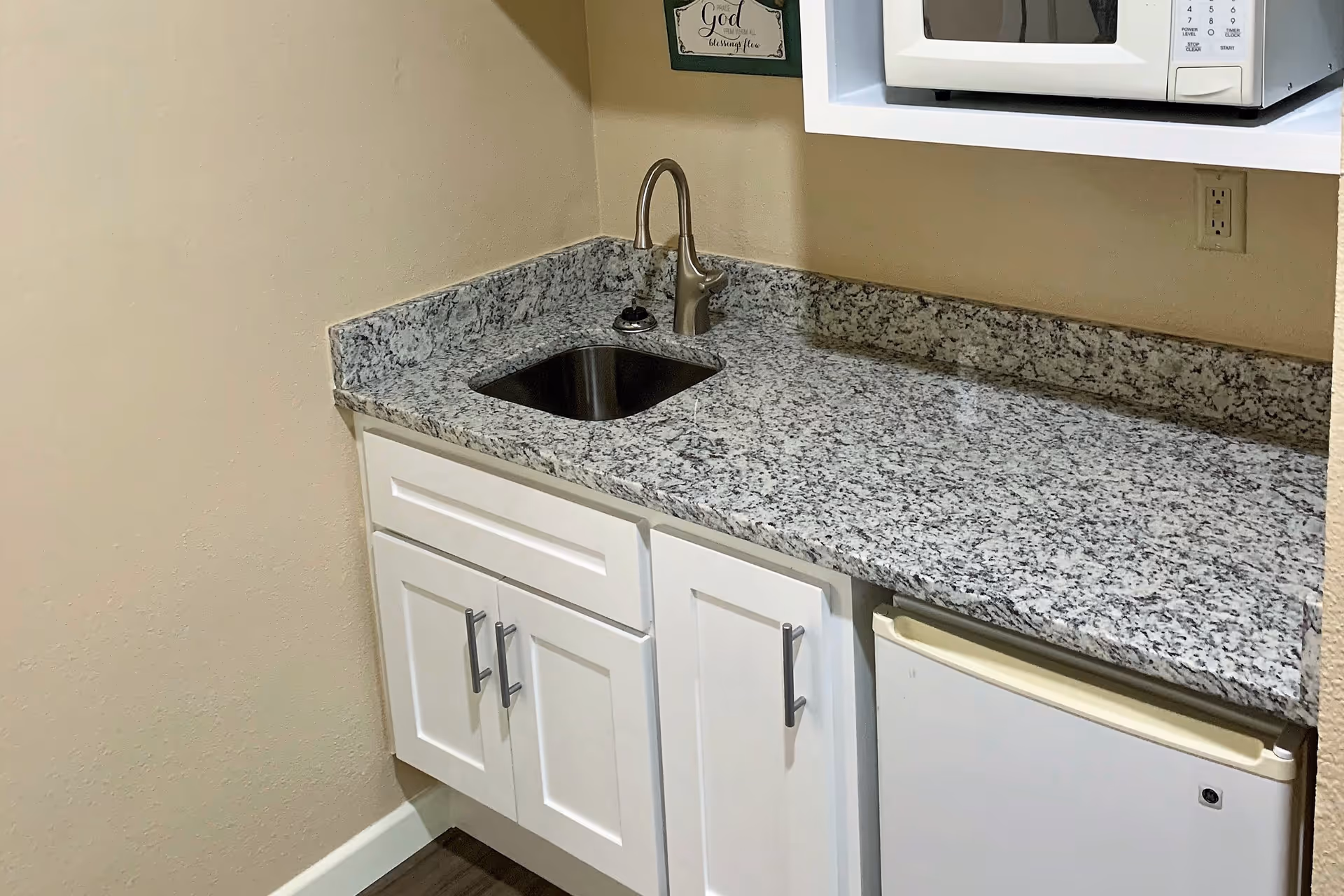 A small kitchen area with a granite countertop, a stainless steel sink with a faucet, white cabinets below, a mini refrigerator, and a white microwave mounted above the counter. A decorative sign is hung on the wall above the sink.