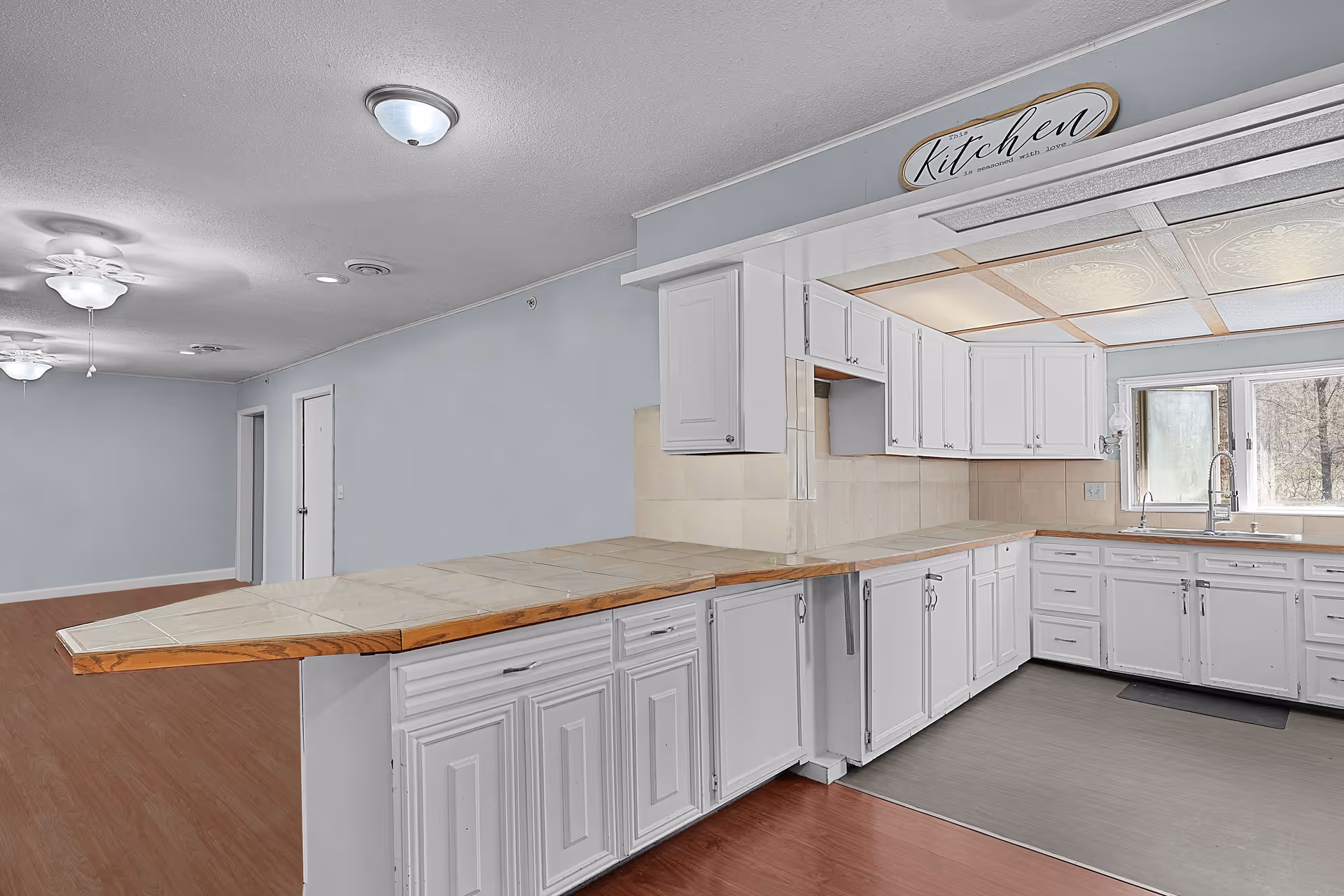 Open white kitchen with a long tiled peninsula, white cabinets, and a sink under a window opening to a large multipurpose room.