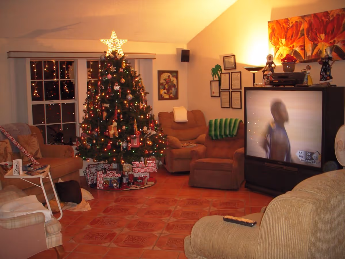 A cozy living room decorated for Christmas with a lit Christmas tree adorned with ornaments and a star on top. Wrapped presents are placed under the tree. The room has several cushioned chairs and a large TV showing a blurred image of a person. The walls have framed pictures and colorful artwork, and the floor is tiled.