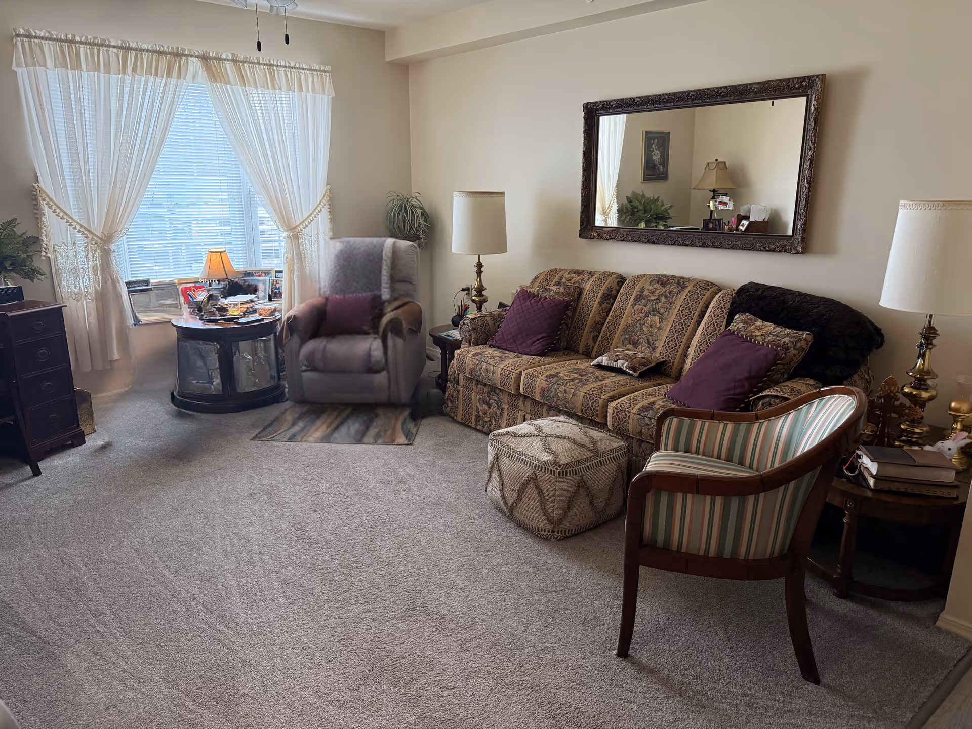 Well-lit living room with a patterned sofa, armchairs, side tables, lamps and a window with sheer curtains.