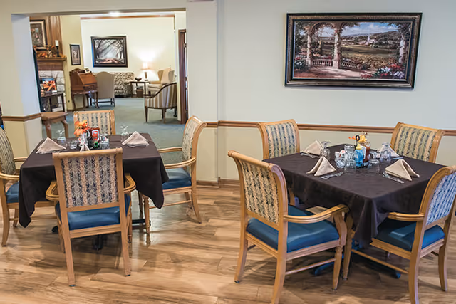 Two dining tables set with black tablecloths, napkins, and condiments in a senior living facility dining area. Each table is surrounded by wooden chairs with blue cushions. In the background, there is a living room area with armchairs, a piano, and framed artwork on the walls.