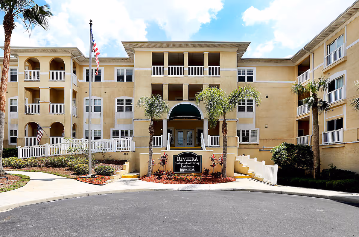 Exterior front view of a three-story beige senior living residence building with balconies, palm trees, an American flag, and a sign that reads 'Riviera Independent Living Residences'.
