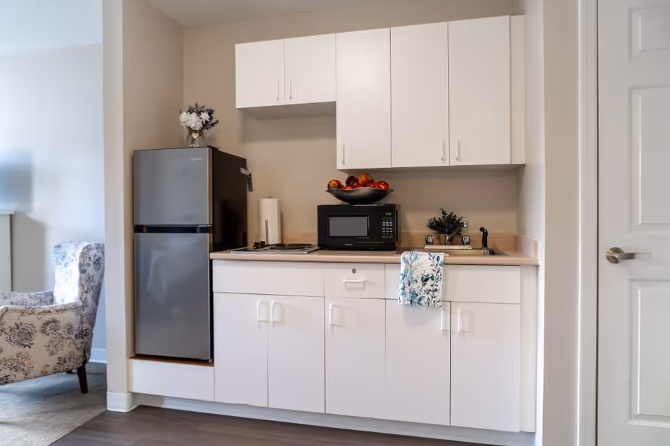 A compact kitchen area with white cabinets, a small refrigerator, a microwave, a sink, and a stovetop. A bowl of apples and a paper towel holder are on the countertop. A floral-patterned towel hangs from the cabinet handle. To the left, part of a floral armchair and a lamp are visible in an adjoining room.