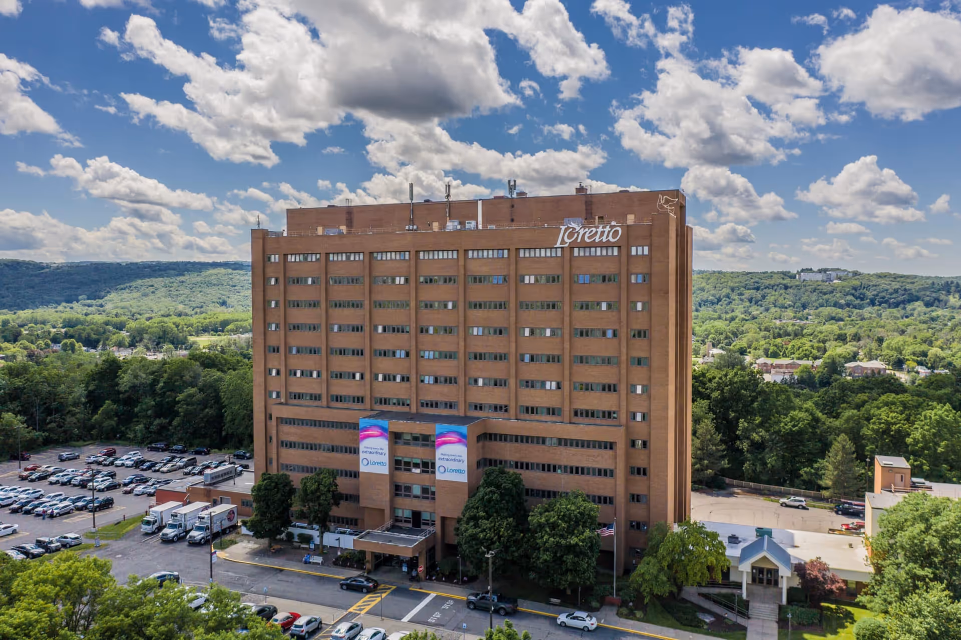 An aerial view of a tall brick building with banners at the entrance, a parking lot, and surrounding green hills under a blue sky.