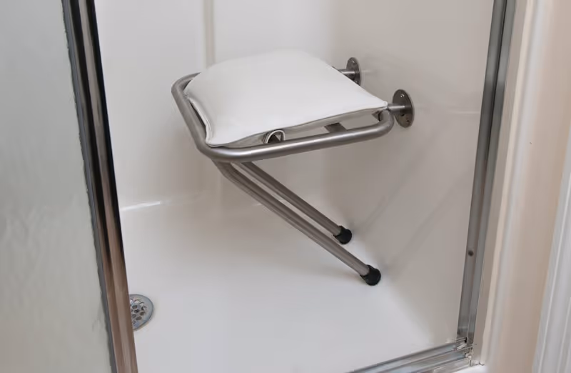 A white fold-down shower seat with a padded cushion mounted on the wall inside a shower stall with a metal frame and a drain on the floor.