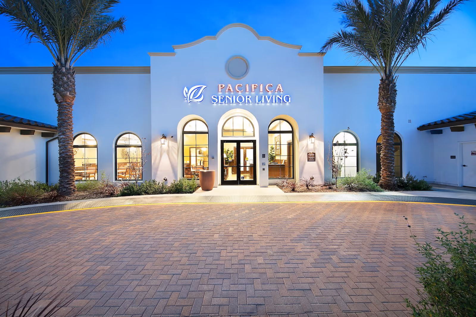 Front exterior view of a senior living facility named Pacifica Senior Living, featuring a white building with arched windows and doors, two tall palm trees on either side, and a paved driveway in front under a clear blue sky.