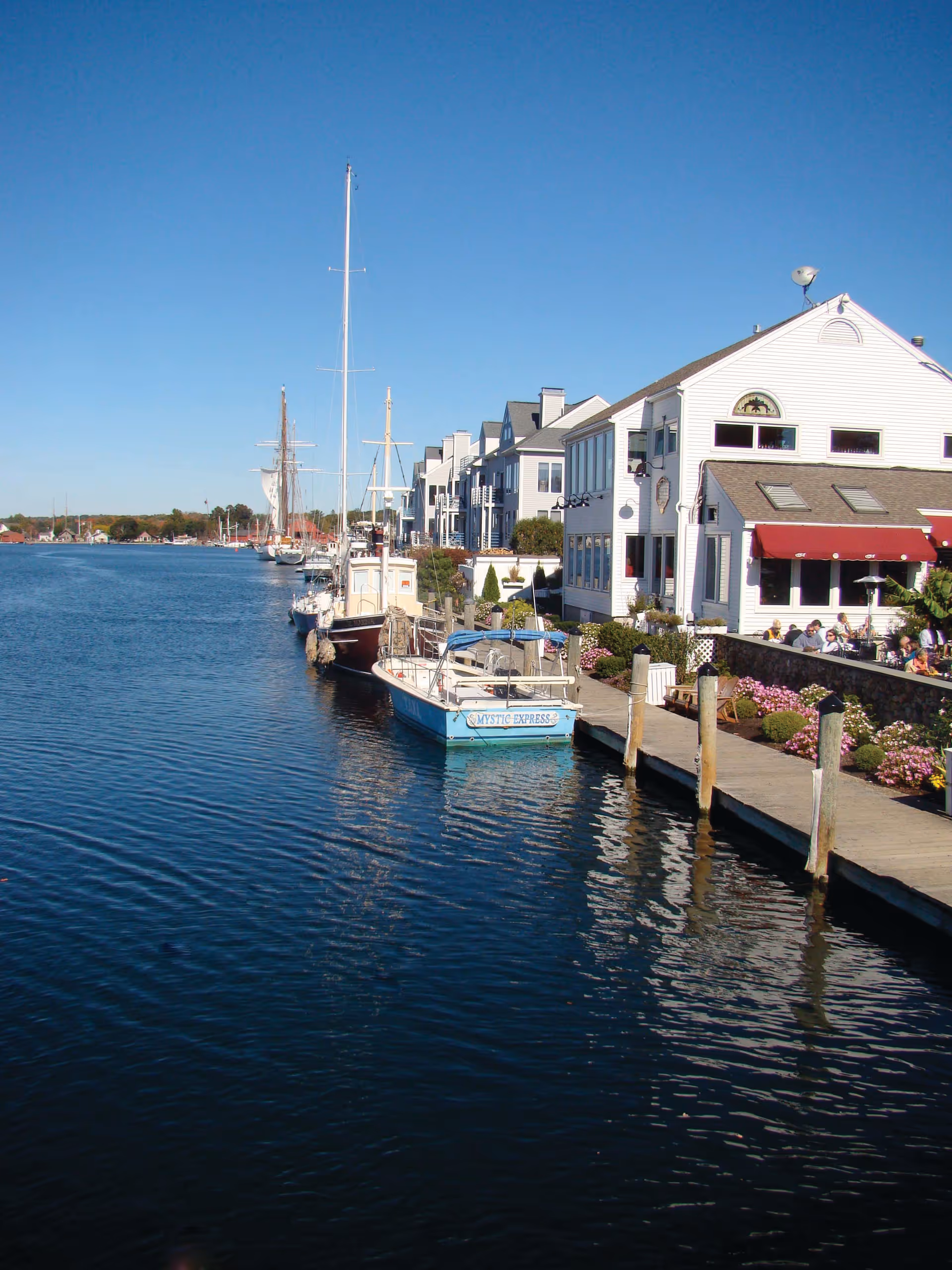 A waterfront scene showing several boats docked along a wooden pier next to white buildings with red awnings. People are sitting outside near the water, and the sky is clear and blue.
