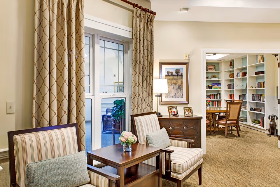 A cozy interior room with two striped armchairs and a small wooden table with a vase of flowers between them. Behind the chairs is a window with patterned curtains. To the right, there is a wooden cabinet with framed photos and a lamp, and an open doorway leading to a room with bookshelves filled with books and decorative items, along with a wooden table and chairs.