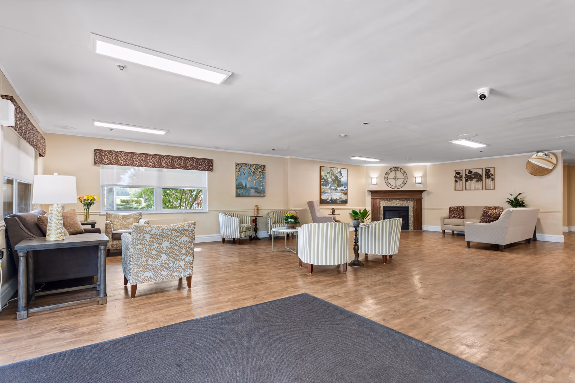 Spacious and well-lit living room area in Batavia Nursing Care Center featuring multiple seating arrangements with armchairs and sofas, a fireplace with a large clock above it, wall art, and large windows with valances allowing natural light to fill the room. The floor is wooden, and there are plants and lamps adding to the cozy atmosphere.