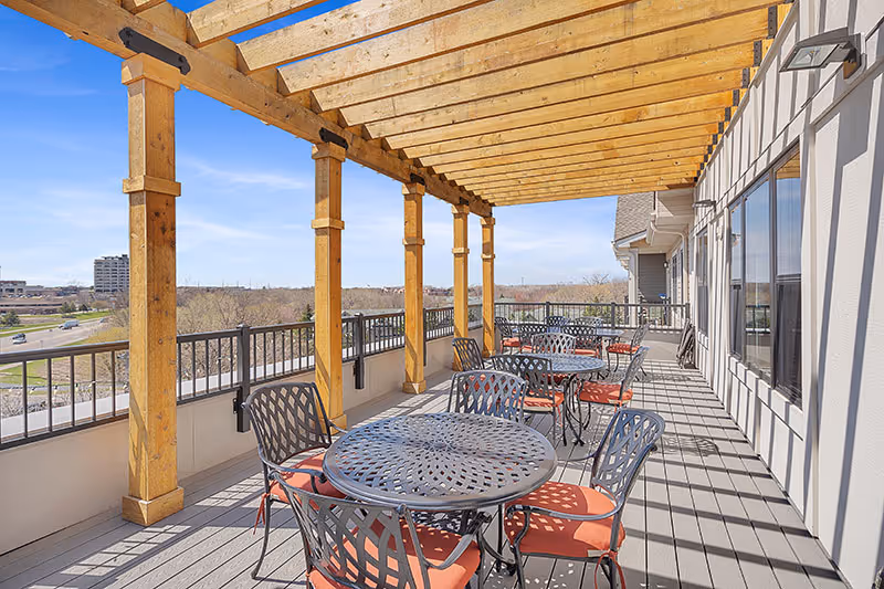 Outdoor patio area with multiple round metal tables and chairs with orange cushions under a wooden pergola. The patio overlooks a scenic view with trees and buildings in the distance under a clear blue sky.