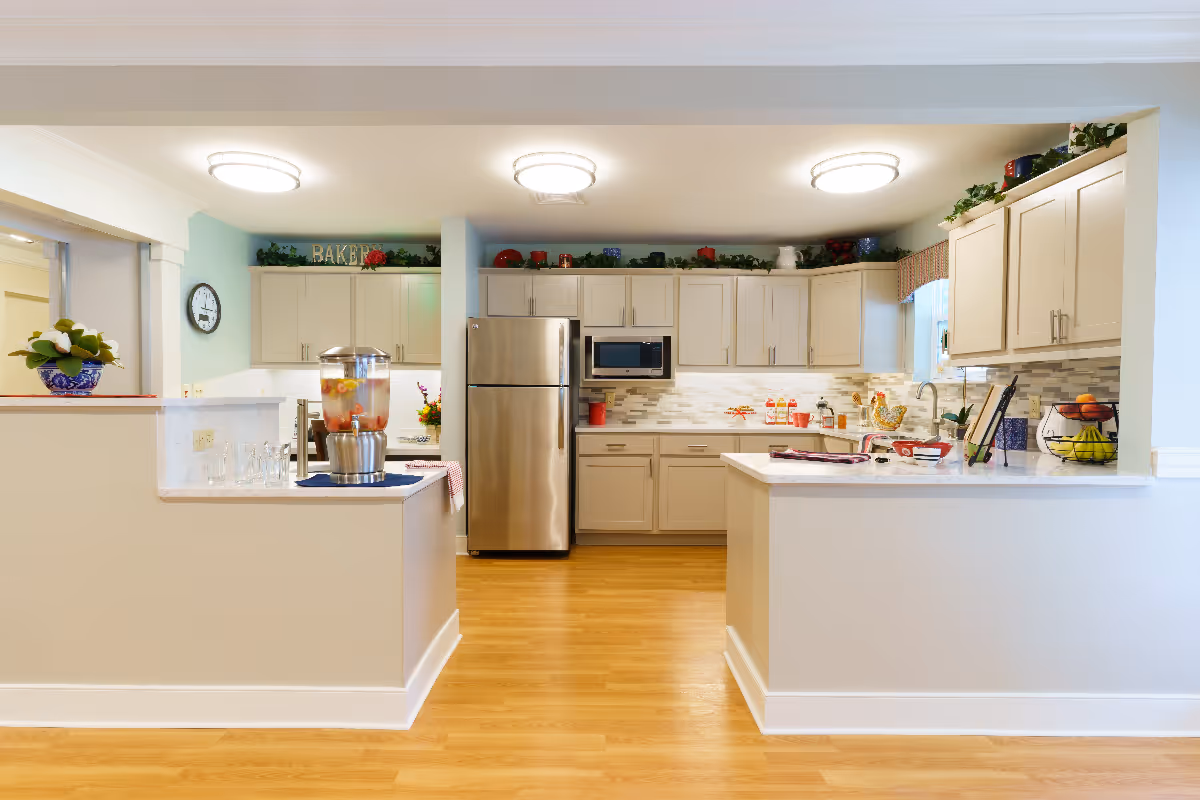A bright and spacious kitchen area with light-colored cabinets, stainless steel refrigerator, microwave, and a countertop with various decorative items and a beverage dispenser. The kitchen has wooden flooring and is well-lit with ceiling lights.