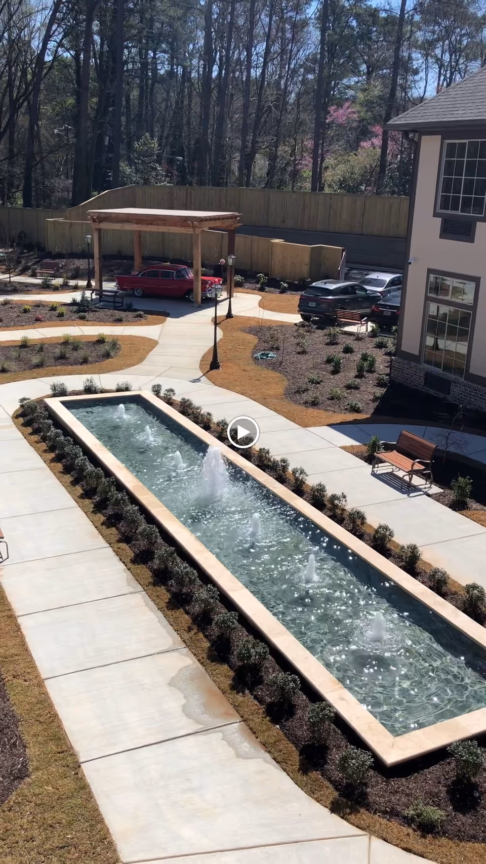 Sunlit courtyard with a long rectangular fountain with multiple water jets, surrounding walkways, benches, a pergola, parked cars, and a building facade.
