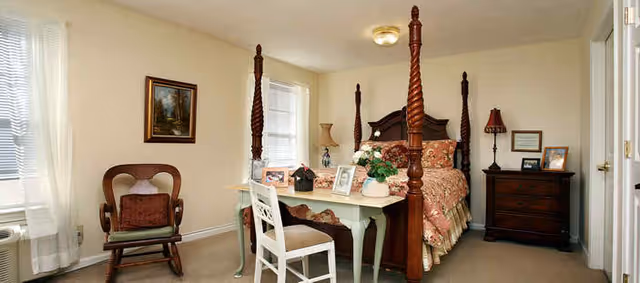 Sunny traditional bedroom with a four-poster bed, small desk and chair, wooden dresser, and an armchair.