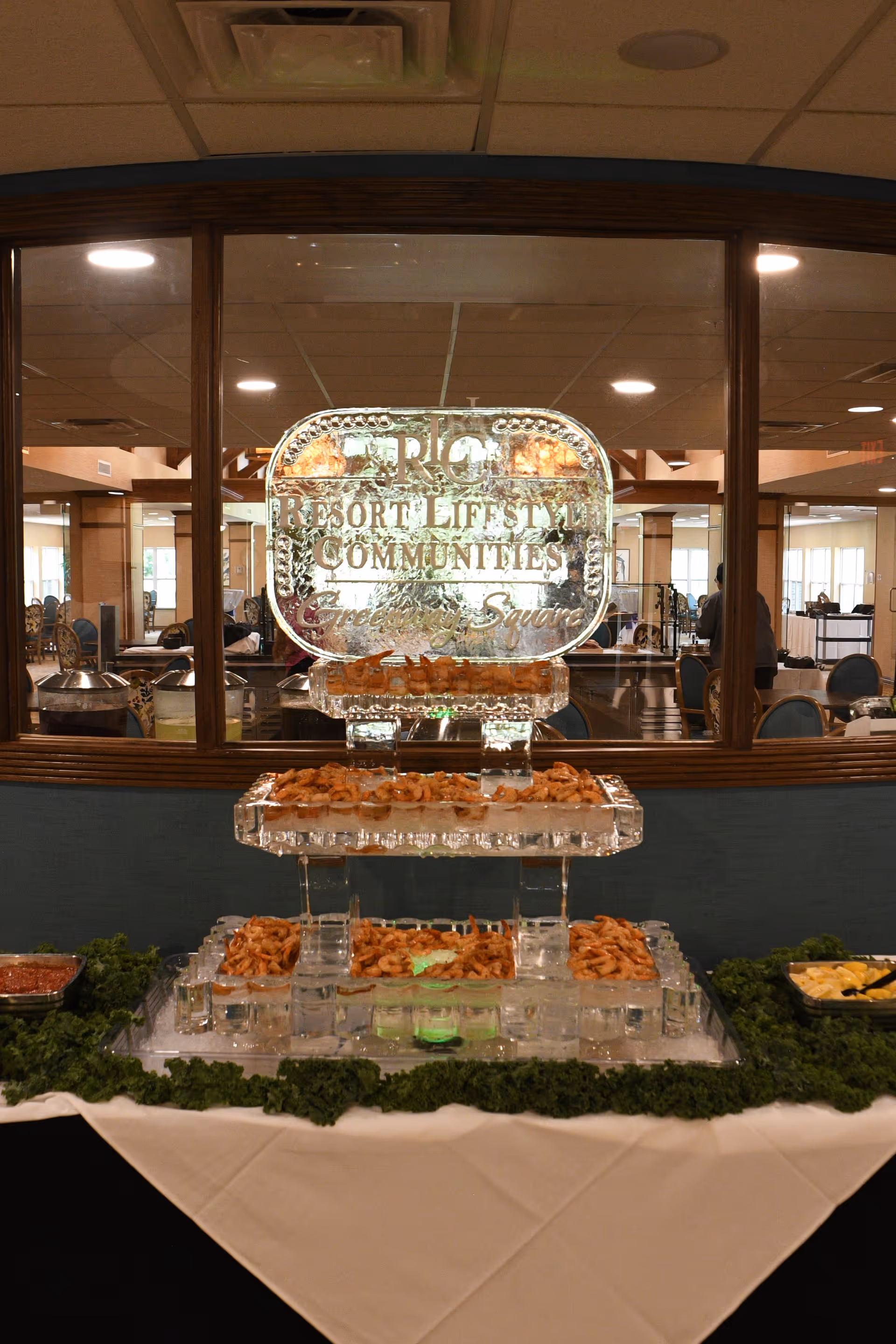 A buffet table with a multi-tiered ice sculpture display holding shrimp, surrounded by greenery garnish. The ice sculpture has the text 'Resort Lifestyle Communities Greenway Square' etched on it. The background shows a dining area with tables and chairs.